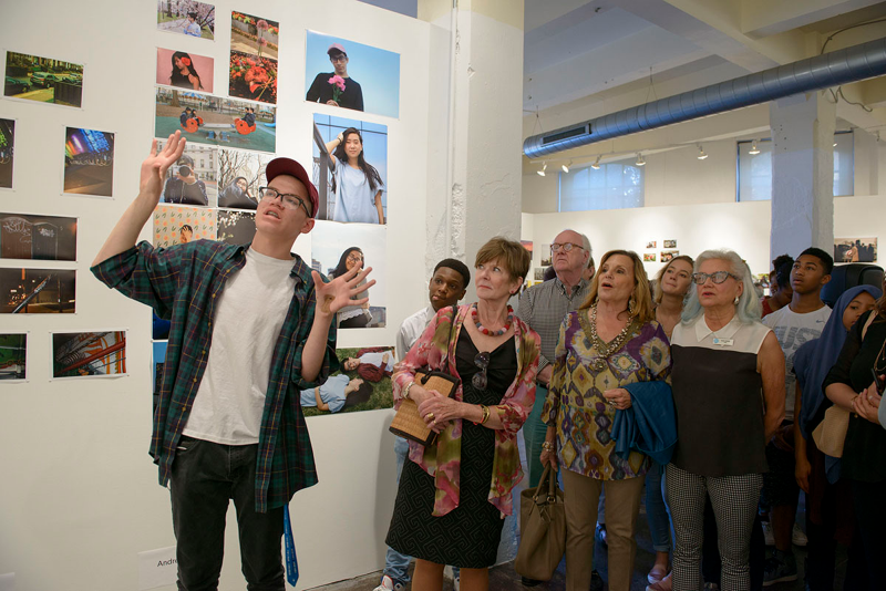 Young person gesturing with their hands and speaking to a crowd about a photo on the wall.