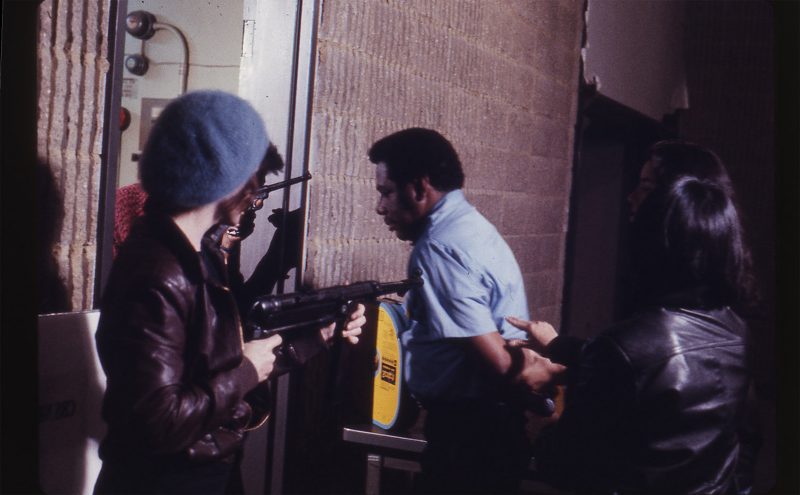 Man being lead into a room with multiple guns pointing at him, from "Born in Flames"