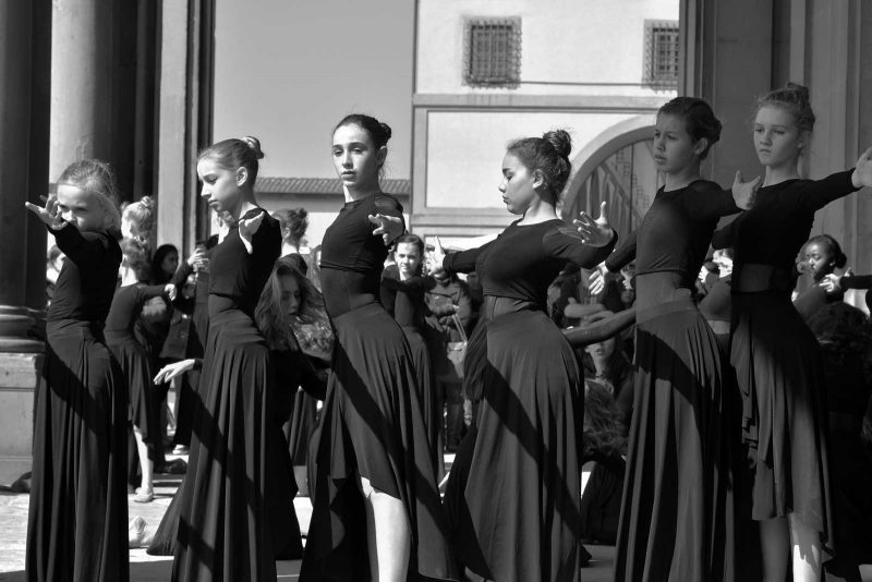 Four girls in long black dresses doing ballet exercises in a straight line outdoors.