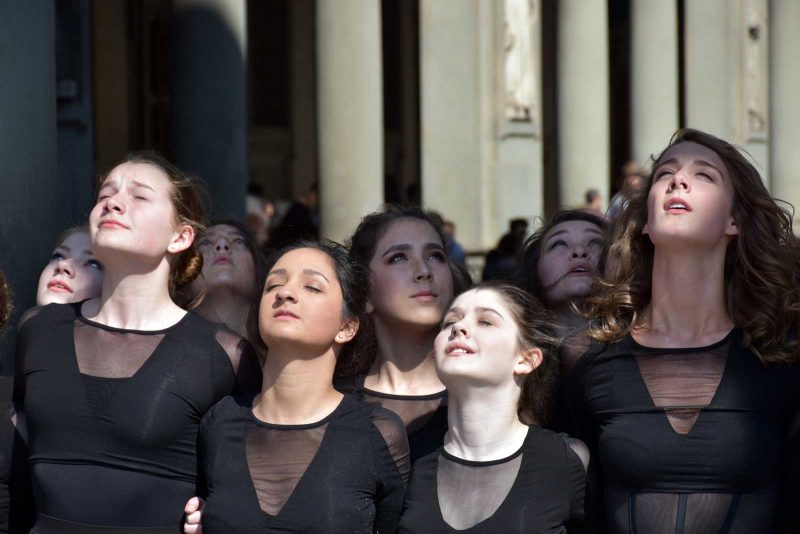 A group of young woman wearing black matching dresses looking upwards towards the sun and holding each other arm in arm.