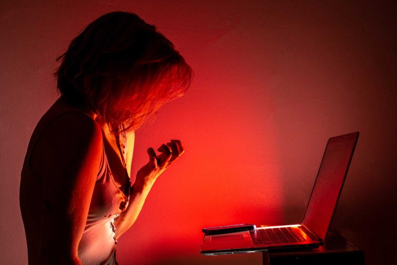 Woman viewed from the site, staring at a computer, consumed by red light, clutching her hand towards her chest.
