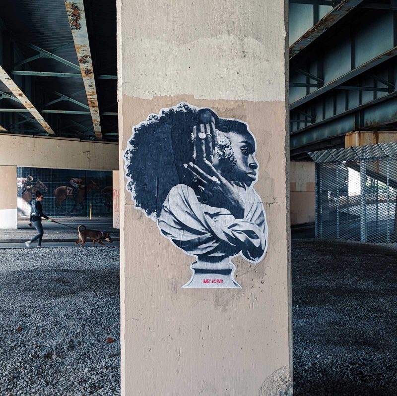 Photo collage of a black woman with long curly hair and her hands covered with rings, covering her ears, pasted onto a parking garage pillar.