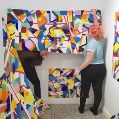 Monica holding up colorful abstract panels and attaching them to the wall.