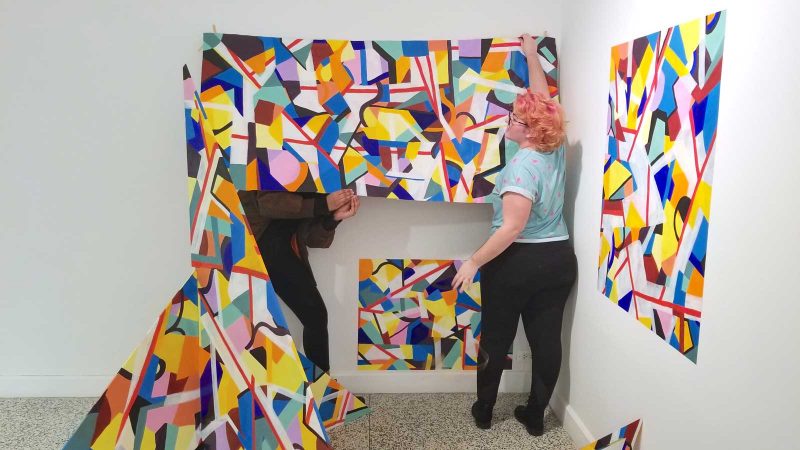Lee holding up colorful abstract panels and attaching them to the wall.