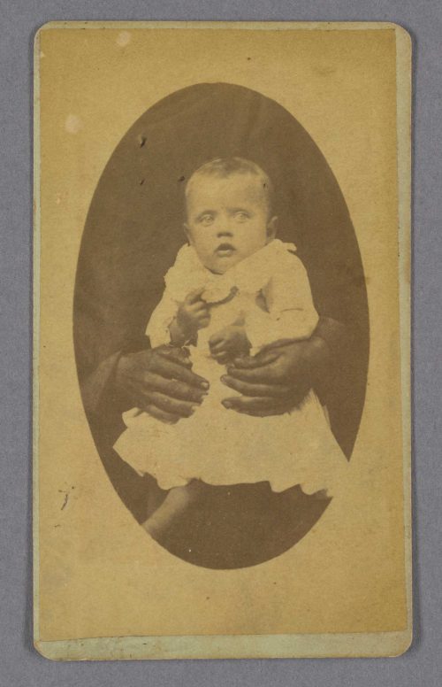 Close-up on a white baby held by a black nanny with only her hands showing.