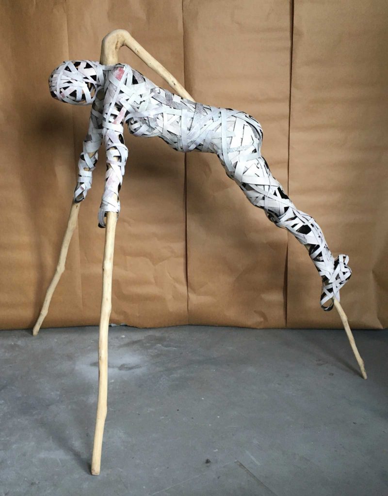 A paper strip sculpture outlining a human body constructed on the structure of a three pronged, long piece of wood that is in a triangular shape. 