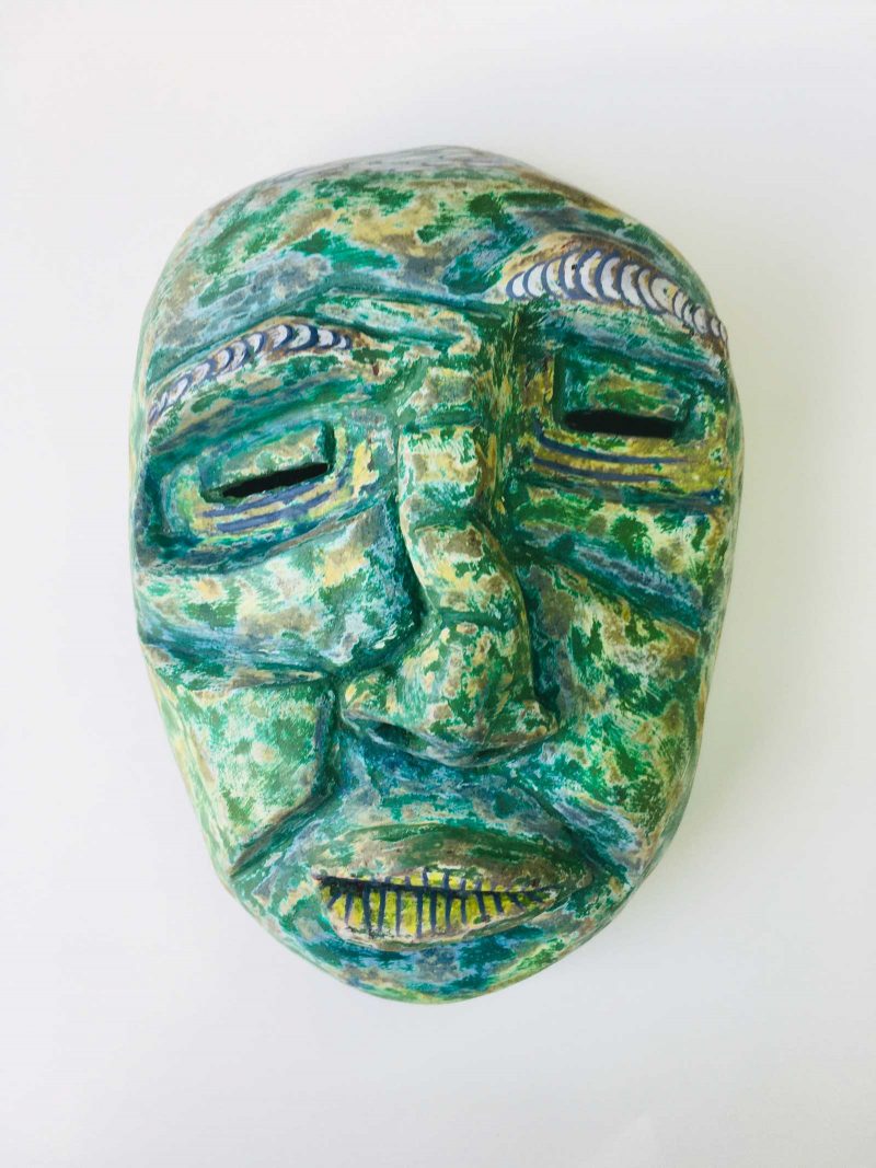 Sculpture of a green mask with crooked features looking downcast and distressed.