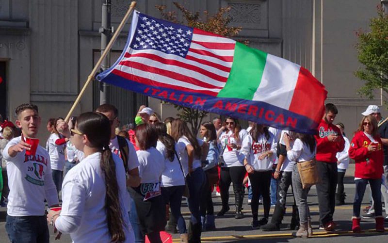 People in white shirts parade through the streets with italian american flags.