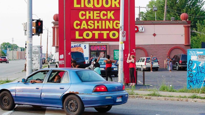 A blue car passing a parking lot with a "Liquor / check / cashing / lotto" sign, people in movement captured below and behind the sign in the lot.