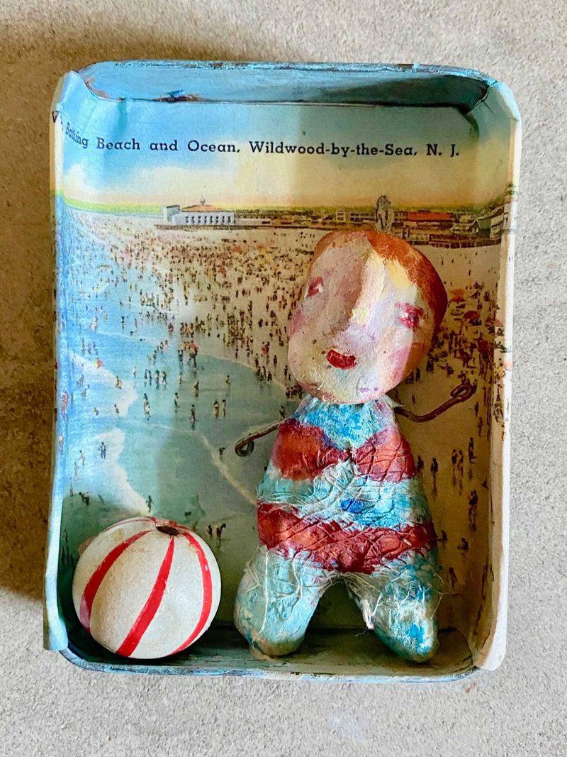 Sculpted female figure in a red bathing suit with a net wrapped around their body sitting in rectangular case with rounded edges. Next to the figure is a white ball with red stripes. Pasted on the case is a beach and the text "Bthing Beach and Ocean, Wildwood-by-the-Sea, N.J."