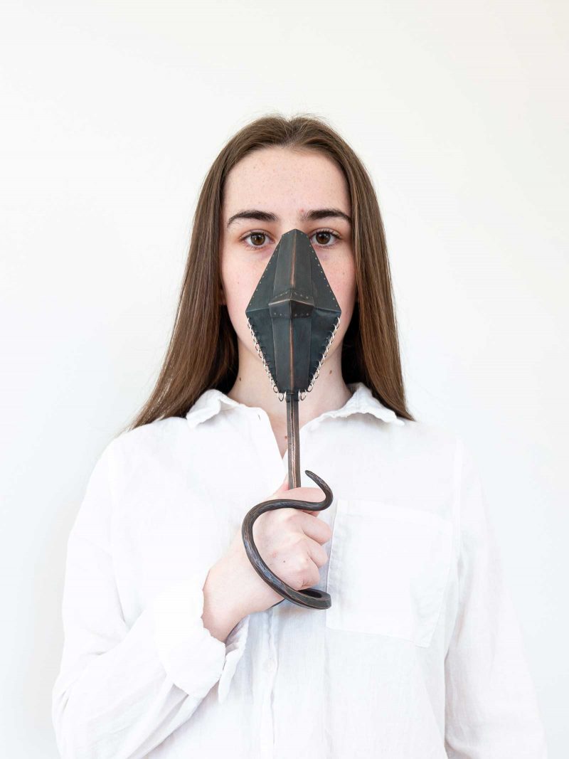 Petite woman holding a sculpture of a kite shaped metal piece with a metal handle which wraps around the woman's hand like a snake.