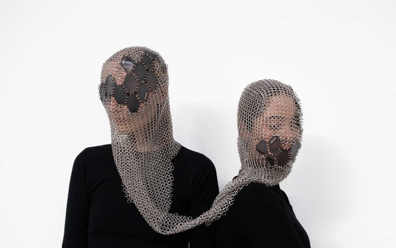 Two women wearing a mesh garment over their heads with metal pentagons woven in to conceal their features that is connected.
