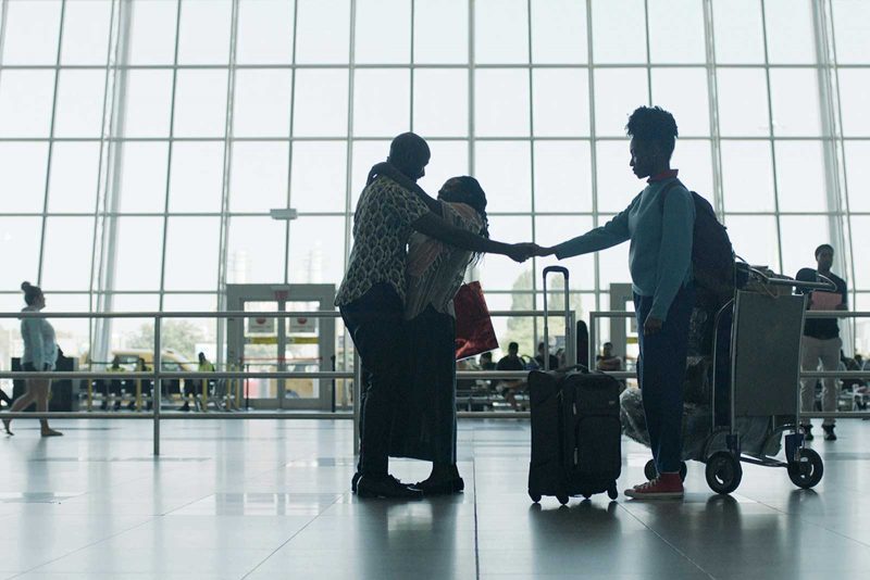 A couple hugging at an airport with their luggage nearby; one of the two embracing outstretches their hand and holds the hand of another person, who is standing nearby.