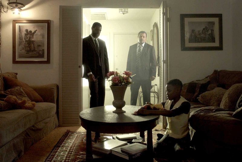 Two grown men stand in a doorway, one partially obstructed by bright light, and look at a young boy sitting on the ground In a living room and playing with something on the coffee table.