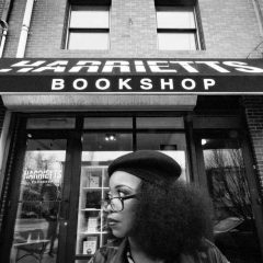 Jeannine A. Cook wearing a beret and glasses looking to the right standing in front of the storefront that says "Harriet's Bookshop"