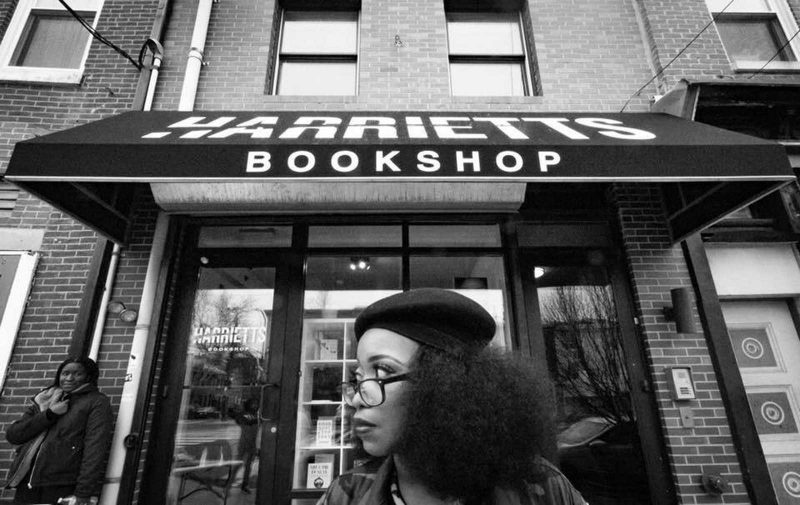 Jeannine A. Cook wearing a beret and glasses looking to the right standing in front of the storefront that says "Harriet's Bookshop"