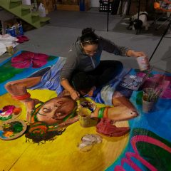 Salina sitting on top of the mural on the ground painting features with one hand and holding paint in the other.