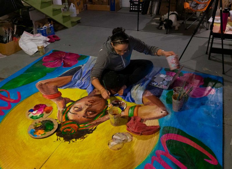 Salina sitting on top of the mural on the ground painting features with one hand and holding paint in the other. 