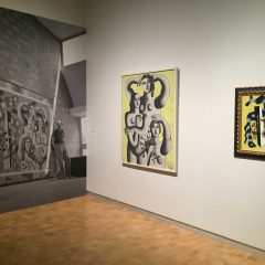 View of the exhibition with a black and white print of pieces on the wall with the same piece framed and shown on the adjacent wall.