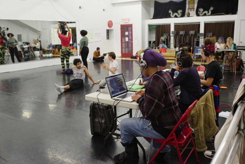 People stretching and warming up in a dance studio and others sitting at a table with headphones and laptops working.