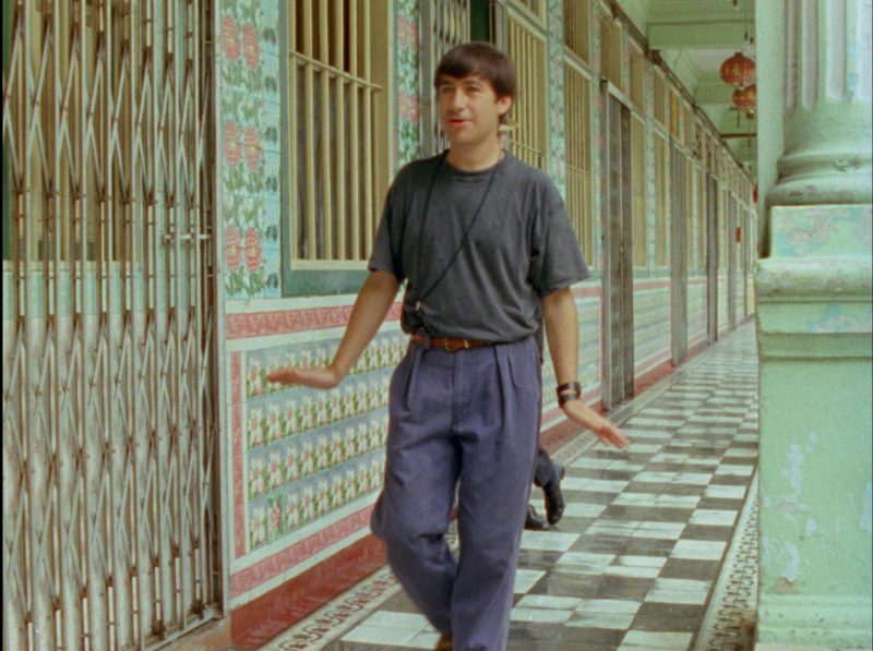 A masculine person wearing a tucked in shirt and pleated pants walks down a heavily patterned decorative hallway with bars on the windows
