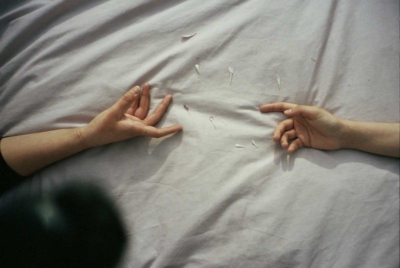 two hands resting on a bed with flower petals, reaching out to each other