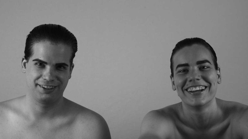 The two bandmembers pictured with bare shoulders from the collarbone up with slicked back hair. They laugh in front of the gray background.