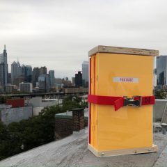 Added Velocity Year Two, More money for five Velocity Fund awardees 4 Stack of zines in a wooden case with a red ratchet strap securing them into a bundle on the edge of a roof with Philadelphia visible in the back.