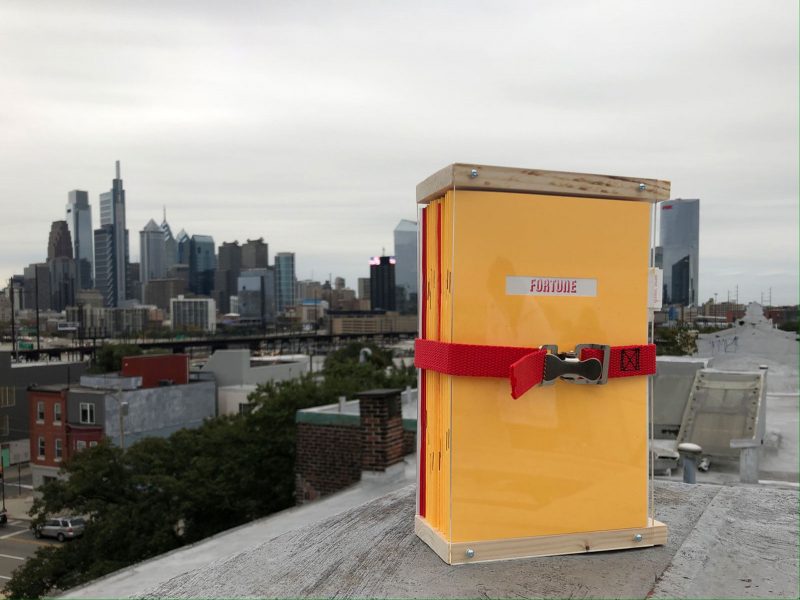 Stack of zines in a wooden case with a red ratchet strap securing them into a bundle on the edge of a roof with Philadelphia visible in the back.