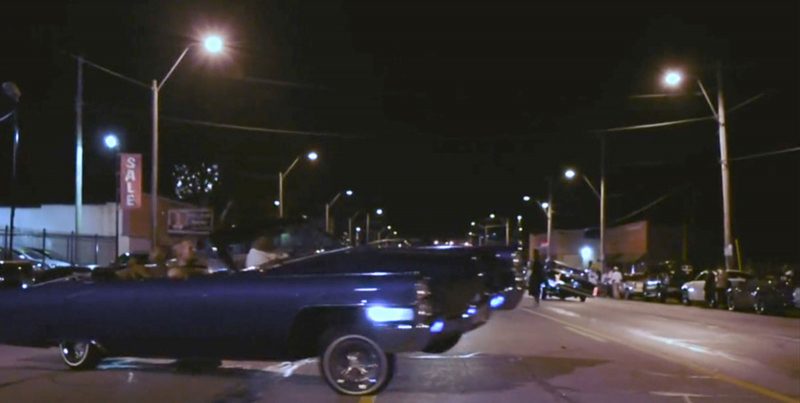Video still of an old fashioned convertible car driving out of a parking lot onto a deserted road.