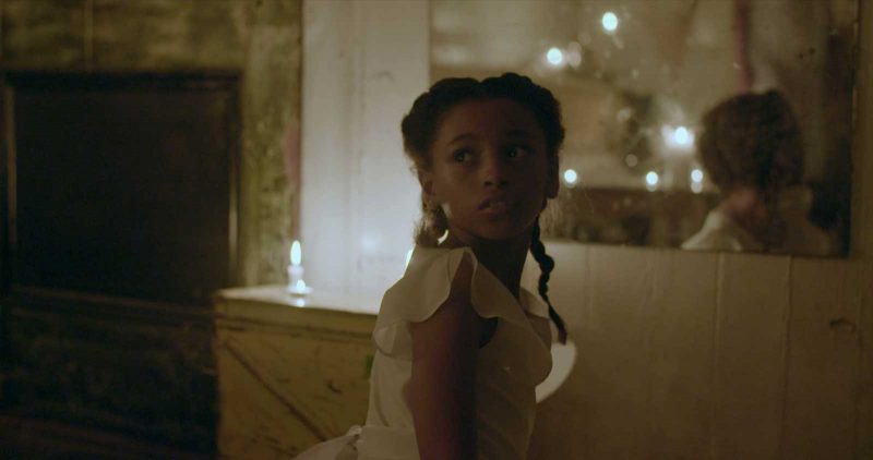 A girl in a white dress who is standing in a low lit room looks over her shoulder at an unknown object or person.