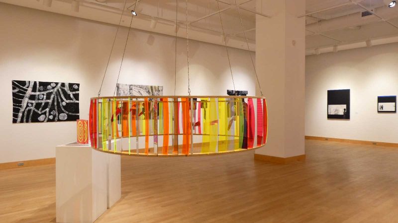 Installation view featuring a large circular sculpture of transparent plastic sheets formed into a wheel shape and hanging from the ceiling by chains.