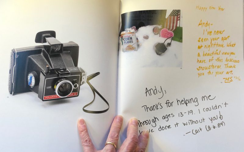 Open book with hand holding book open. Left page shows Polaroid camera, right page shows text and a snapshot of a grave.