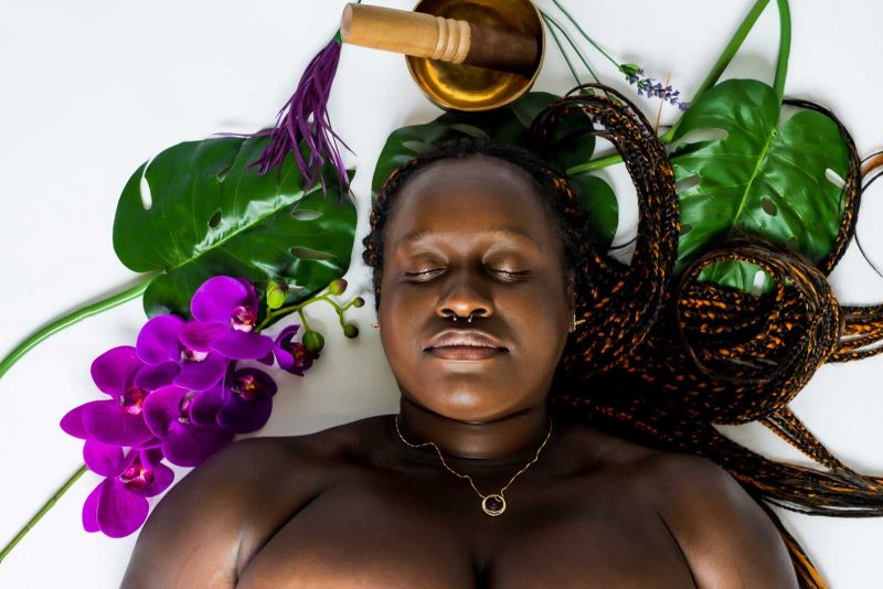 A black person with long black and orange braided hair, wearing a gold necklace, laying on a white background that is decorated with purple flowers (which sit to the right of the figure's shoulder), two large green leaves (one to the left and one to the right of their head), and a golden bowl with a wooden cylinder sitting inside of it (above the figure's head).