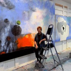 Diane Burko in her studio, leaning against a step stool and standing in front of a large subtract landscape painting of a blue atmospheric surface with abstracted planets on the bottom left, next to a bright orange explosion of color, both coming out of a black patch at the bottom of the canvas.