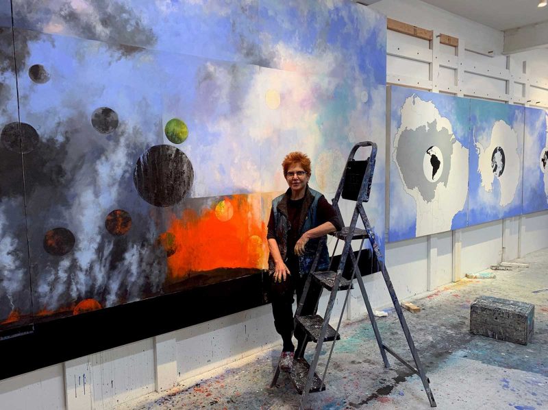 Diane Burko in her studio, leaning against a step stool and standing in front of a large subtract landscape painting of a blue atmospheric surface with abstracted planets on the bottom left, next to a bright orange explosion of color, both coming out of a black patch at the bottom of the canvas.