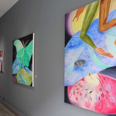 Gray gallery wall with two large canvases and one small canvases, all of which have dark backgrounds with colorful, pastel colored imagery of butterflies and flowers. One of the large paintings (on the far right) also includes two legs visible only from below the pelvis down to the feet, floating from the top of the canvas and extending to the middle.