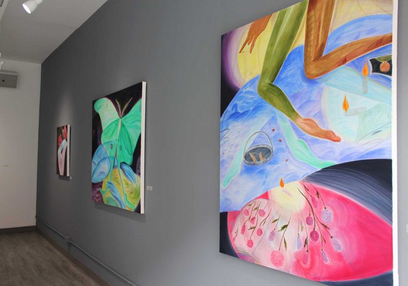 Gray gallery wall with two large canvases and one small canvases, all of which have dark backgrounds with colorful, pastel colored imagery of butterflies and flowers. One of the large paintings (on the far right) also includes two legs visible only from below the pelvis down to the feet, floating from the top of the canvas and extending to the middle.