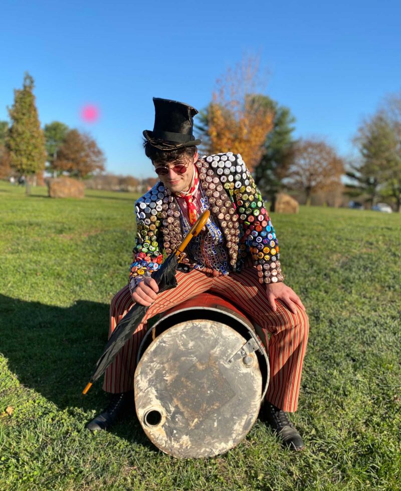 A white man, wearing a carnival inspired costume fashioned out of recycled materials, grins and sits on an old oil drum. He is performing as character "The Barker."