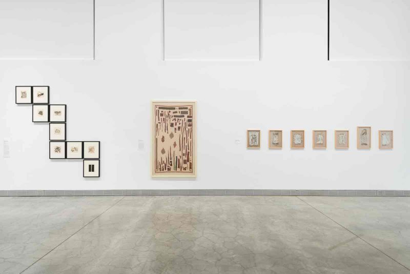 An exhibition view of “Fault Lines: Contemporary Abstraction by Artists from South Asia” at the Philadelphia Museum of Art. 17 framed artworks of various sizes arranged into 3 sets on one large wall. On the left, 9 medium-sized works on paper sit inside of black frames. They are arranged in a varying zig-zagging shape stretching downwards from the top left to the right. In the middle, there is one large piece that is almost as tall is the zig-zagging installation. To the right of that is a series of 8 small works on paper hung in a horizontal line.