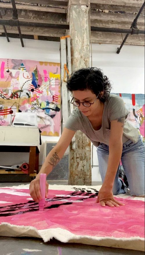 Jackie Milad standing over a piece of canvas and drawing onto it with a marker. Behind her, more of her paintings are hung up on tall, white walls. 