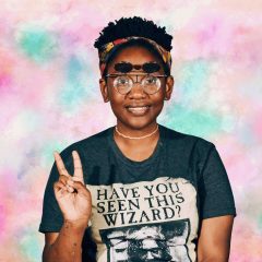 Keyonna Butler, a black woman with short curly black hair who is wearing a colorful headband, retro flip-up glasses, a gray Harry Potter shirt that says "Have you seen this wizard?", and a pink choker necklace. She is smiling and making a peace sign with two fingers with her right hand. There is some paint on her right forearm. The background is a digitally rendered, pastel colored, tie-dye pattern.