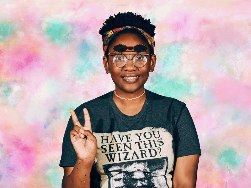 Keyonna Butler, a black woman with short curly black hair who is wearing a colorful headband, retro flip-up glasses, a gray Harry Potter shirt that says "Have you seen this wizard?", and a pink choker necklace. She is smiling and making a peace sign with two fingers with her right hand. There is some paint on her right forearm. The background is a digitally rendered, pastel colored, tie-dye pattern.