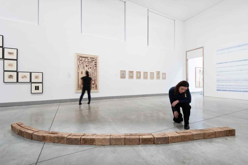 A large exhibition space in the Philadelphia Museum of Art. In the foreground there is an installation on the ground consisting of rectangular brick-like shapes arranged in a slightly curved line. A white person with longish brown hair wearing all black kneels down to look at the sculpture. Behind them, there is a large framed work hanging in the middle of the wall, as well as smaller works arranged in patterns on the left and right of the large work. Another person stands in the back, admiring the large framed work. 