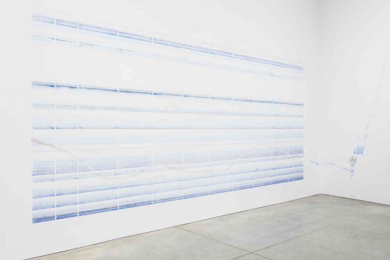 A large fabric artwork hanging on a white wall in the Philadelphia museum of art. The piece takes up the majority of the wall. It has 11 faded blue horizontal lines running across it, each with varying widths and space between the other. There are 10 vertical white lines, disrupting the blue lines. 