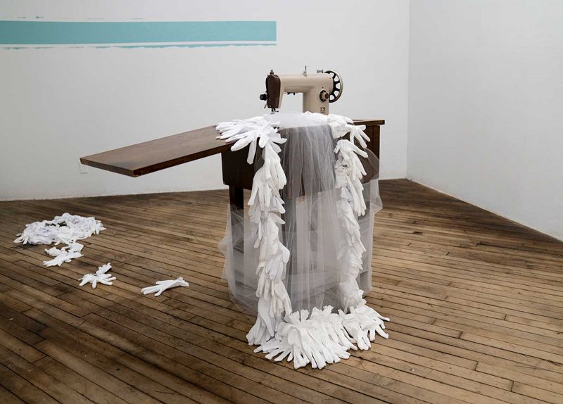 Installation by Imani Roach, of a sewing machine with a sheer garment with fringed edges made out of gloves. 