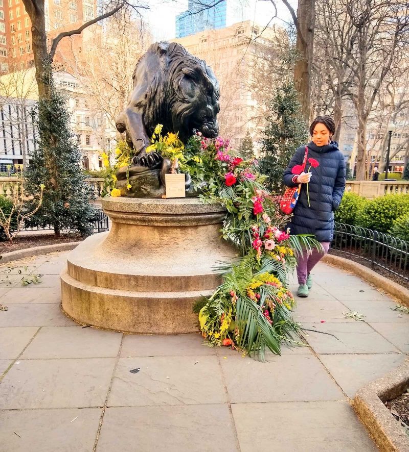 Imani Roach smiling and carrying two flowers past a sculpture and floral arrangement in Rittenhouse Square Park, Philadelphia.