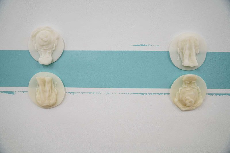 Four wax sculptures by Imani Roach. Each is of the back of a woman's head, showing her hair tied up in a bun. They are installed on a blue horizontal strip of paint on a white wall.