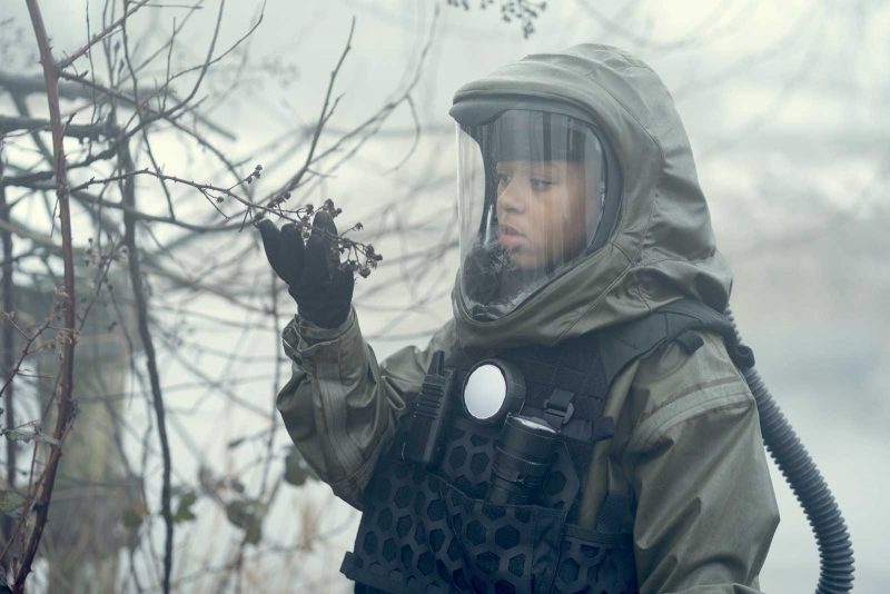 A young Latinx girl wearing a hazmat suit looking wondrously at a tree brand and feeling it with her right hand.