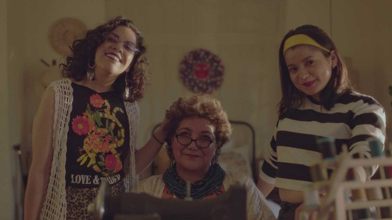 Three Latinx women of varying ages -- two young and one quite old -- gathered in a row. The two young women stand on either side of the older woman, who is sitting. They are smiling and laughing.